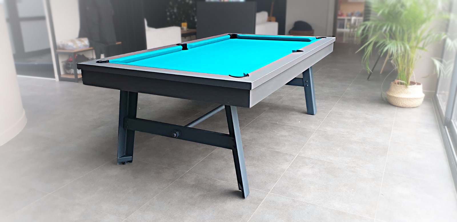 billard vintage en finition nano graphite mat aux pieds laqués anthracite F271 en métal avec tissu vert-bleu carrelage dans un salon au sol en grès cérame émaillé derrière le canapé de la TV à côté de la plante de la baie vitrée qui donne sur la terrasse